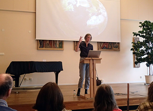 Bilden visar en konferenslokal med talare vid ett podium och en projektorduk bakom som visar jordklotet sett från rymden. 
