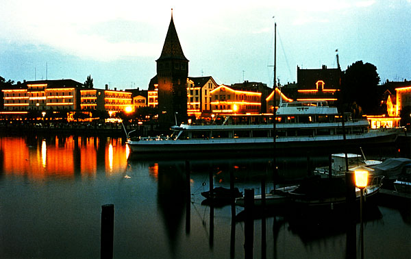 Bilden visar en hamnstad i skymningen, med dekorbelysning på husfasaderna som speglas i vattnet och en passagerarbåt i förgrunden. Passagerarbåten har ungefär samma storlek som en Vaxholmsbåt.