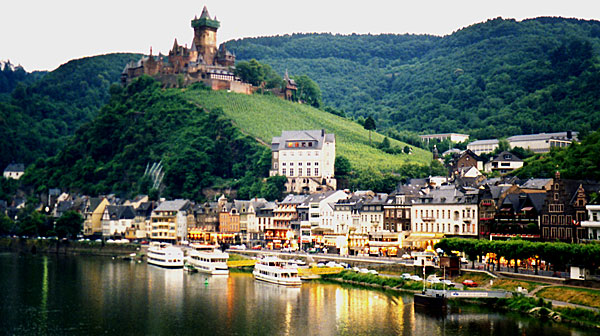 Bilden visar borgen på den spetsiga kullen, med stadsbebyggelse mellan kullen och floden samt tre passagerarbåtar längs flodkanten.