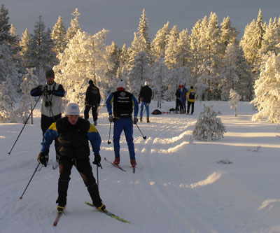 Bilden visar vintrig miljö med långa solskuggor. Några skidåkare värmer upp.