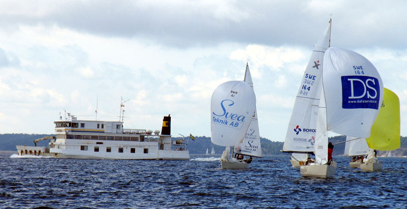 Bilden visar knoX i täten av en grupp Expresser med spinnaker uppe, på väg mot kameran. I bakgrunden passerar en Vaxholmsbåt.