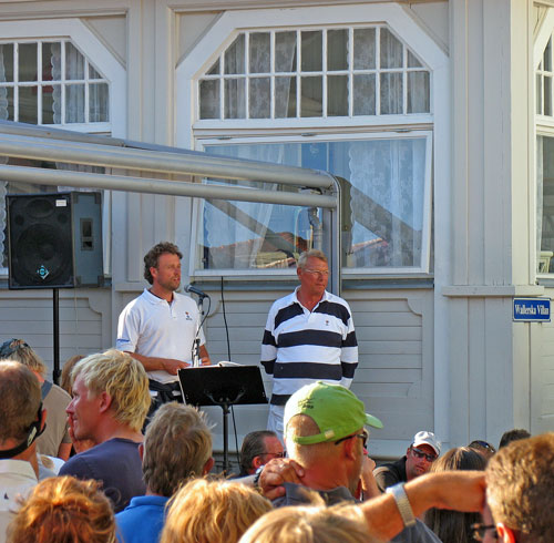Bilden visar två arrangörer som talar i mikrofon. I förgrunden syns många huvuden bakifrån.