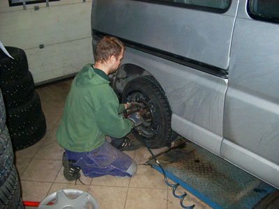 Bilden visar bakhjulet på en minibuss, som står i en däckverkstad. En mekaniker arbetar på huk vid bakhjulet.