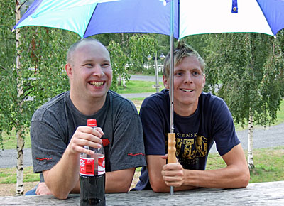 Bilden visar två supportrar under ett golfparaply. De sitter på utemöbler och är glada.
